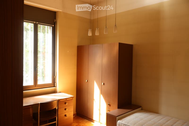 Bright bedroom with wooden floor, large window, bed, wardrobe, desk, and chair.