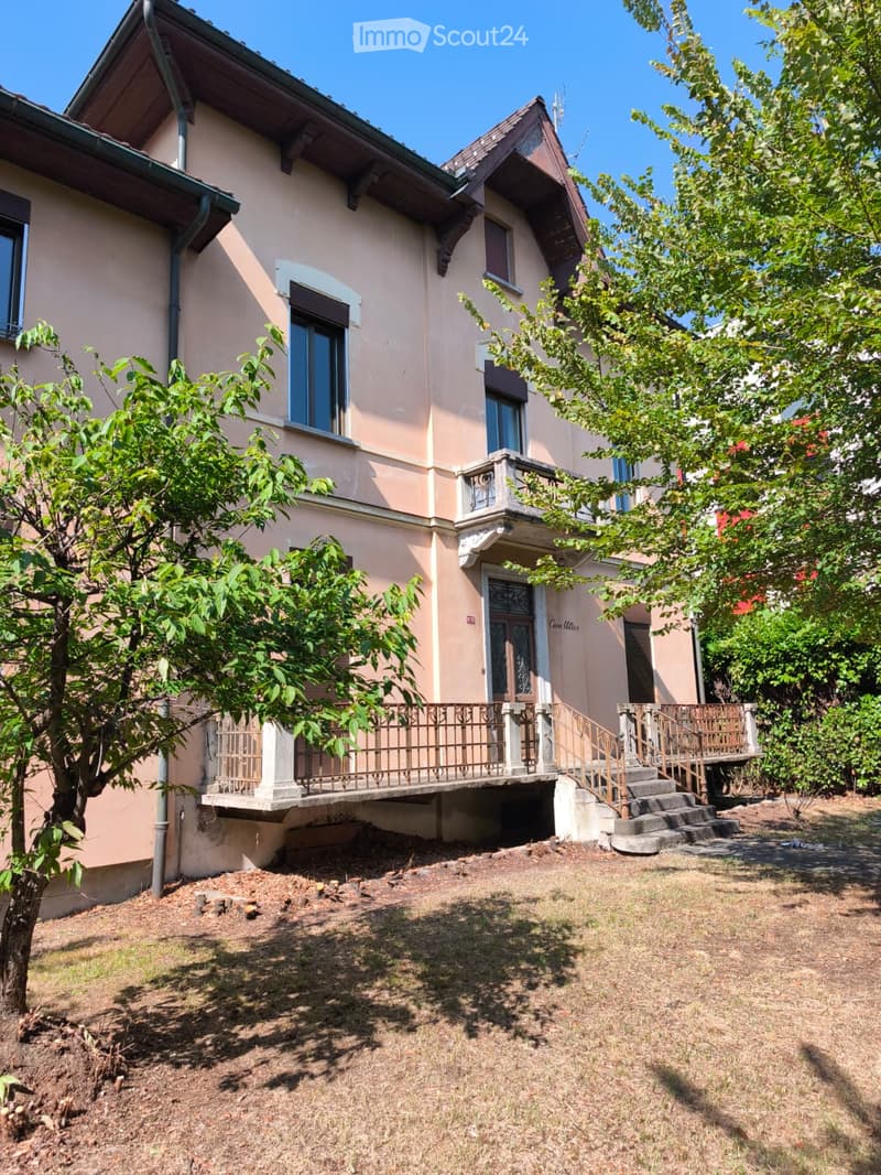 Two-story house with beige walls, multiple windows, and a small balcony with metal railing. The house has a staircase leading up to the entrance and is surrounded by trees and grass.