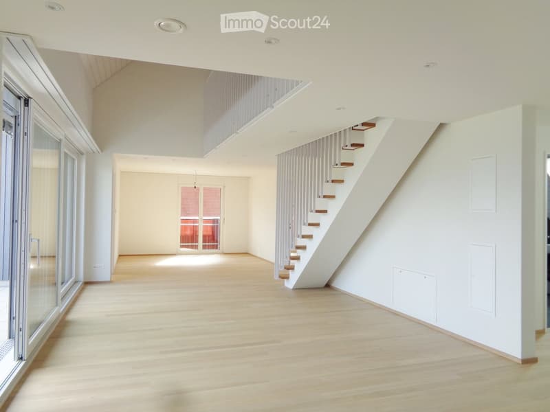 Open, modern interior with light wooden flooring, white walls, and a staircase leading to an upper level. Large windows and glass doors provide natural light.