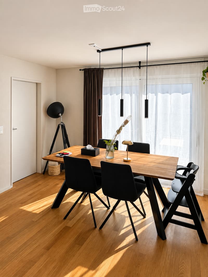 Modern dining area with a wooden table, black chairs, pendant lights, and a decorative vase on the table. The room features light-colored walls, wooden flooring, and large windows with sheer curtains.