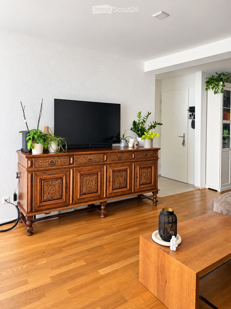 Living room with wooden flooring, a large wooden cabinet with intricate carvings holding a television and several potted plants, a wooden coffee table with a decorative candle holder and small figurine, and a white bookshelf with glass doors in the backgr
