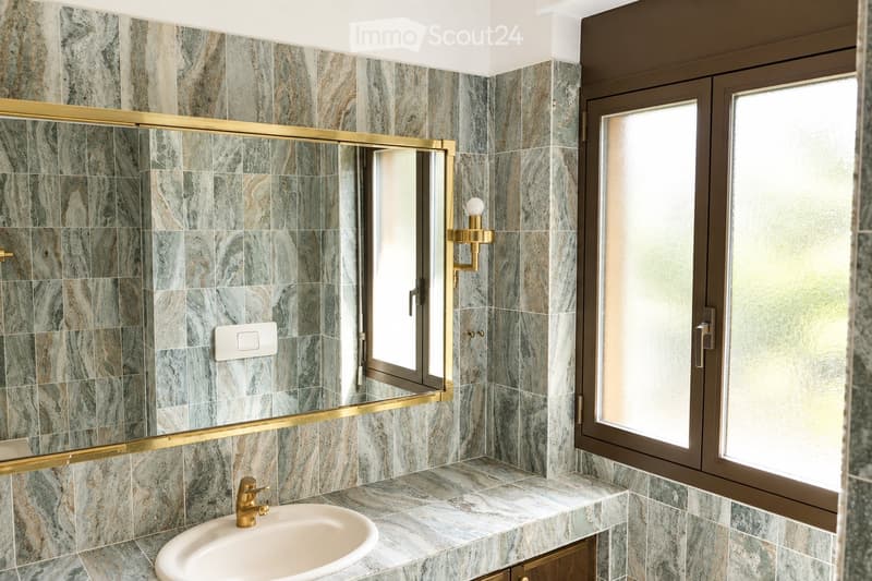 Modern bathroom with marble tiles, a gold-framed mirror, a sink with a gold faucet, and a wall-mounted light fixture.