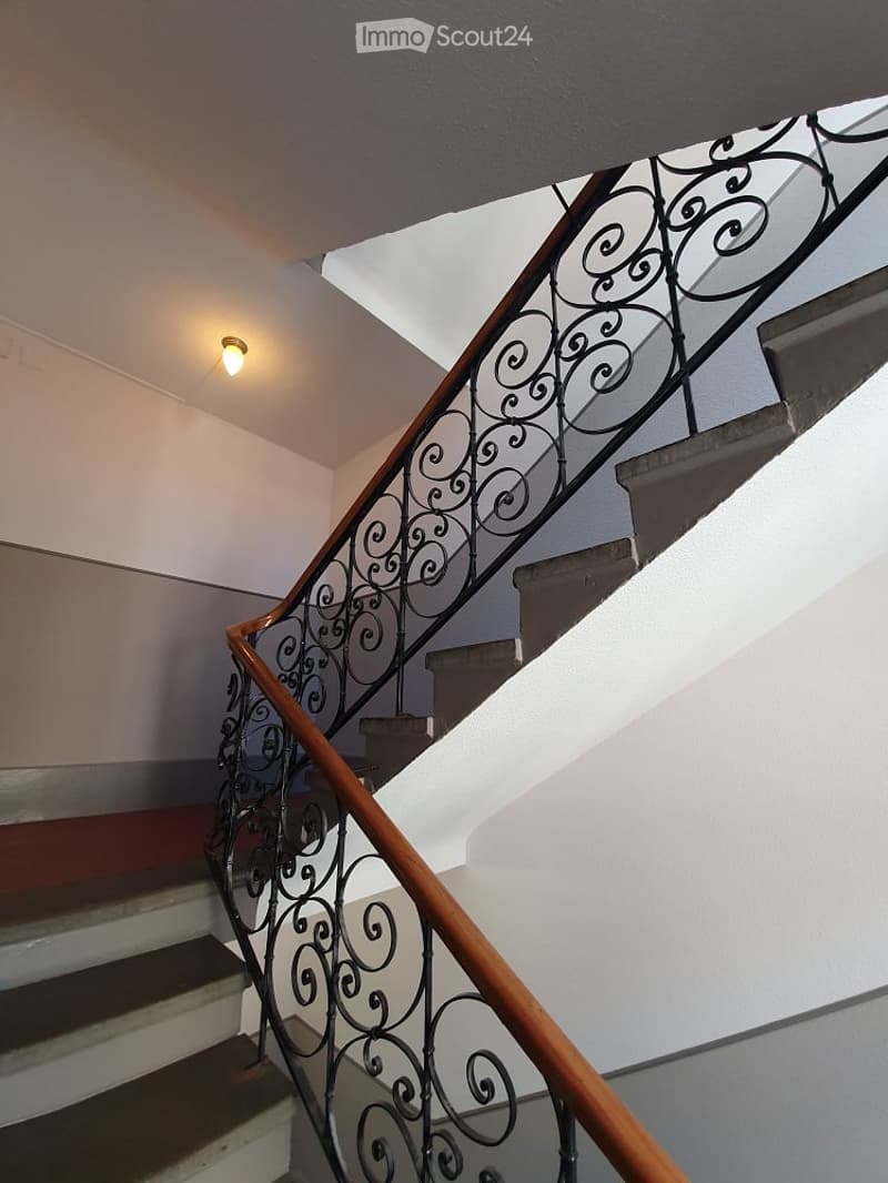 Staircase with wooden handrail and ornate metal balustrade, white walls, and a single ceiling light fixture.