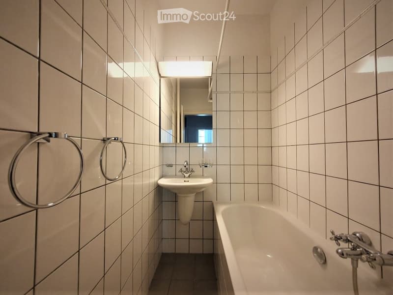 Modern bathroom with white tiled walls and floor, featuring a sink, bathtub, mirror, and towel ring.