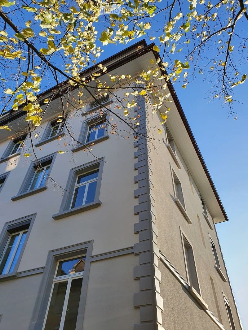 Four-story residential building with light beige facade, multiple windows, and decorative vertical siding. Overhanging roof and tree branches visible in foreground.