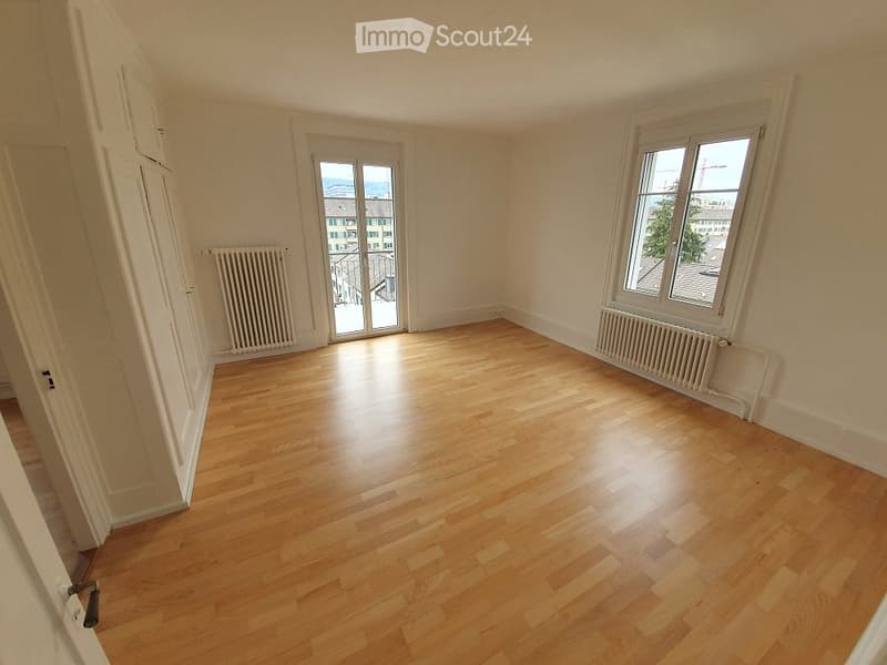 Empty room with wooden flooring, white walls, and large windows offering a view of the cityscape.