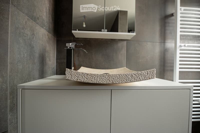 Modern bathroom with a sleek, minimalist design. Features a stone-textured sink, chrome faucet, and a wall-mounted mirror above a white cabinet.