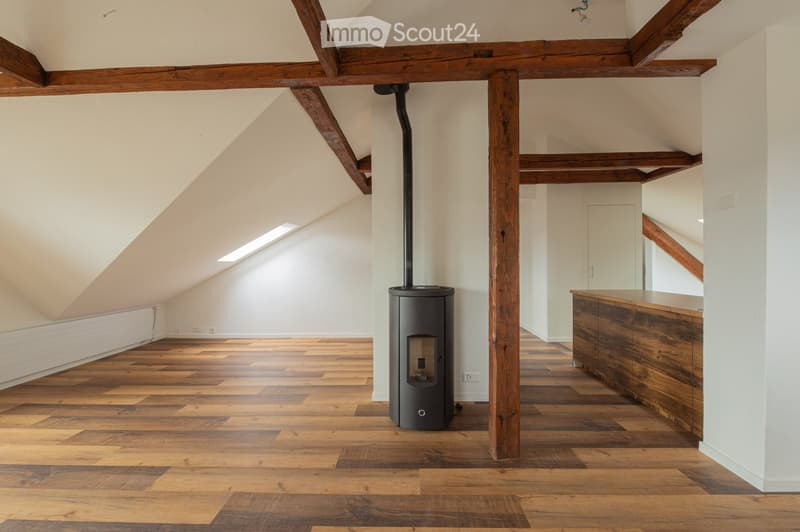 Open loft-style room with wooden beams, polished wooden floor, and a modern black wood-burning stove.