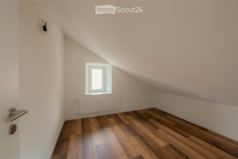 Empty attic room with wooden flooring, white walls, a single window with blinds, and a radiator below it. The ceiling slopes, and there is a door with a silver handle on the left side.