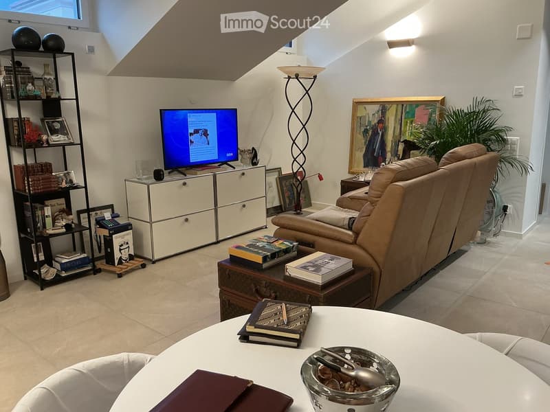 Modern living room with a beige sofa, coffee table, books, television, shelf, and a floor lamp. The room features a slanted ceiling and large windows, creating a bright and airy atmosphere.