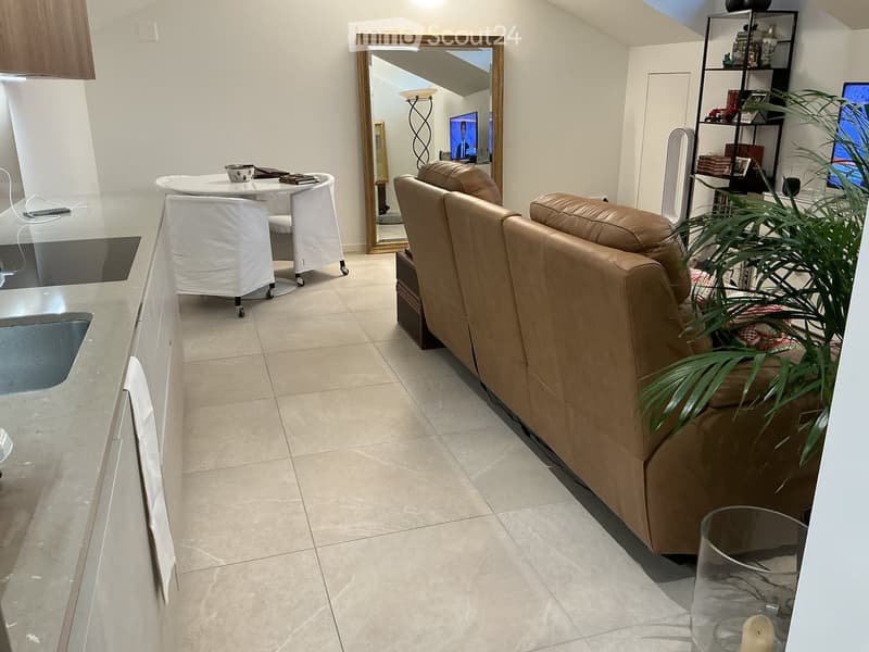 Modern living room with a beige leather sofa, a round white table with chairs, a large mirror, a bookshelf, and a potted plant. The room features tiled flooring and a sleek kitchen area with a glass countertop.