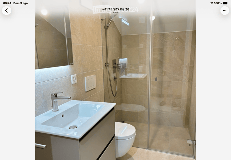 Modern bathroom with beige tiles, a white sink with a chrome faucet, a large mirror, a toilet, and a glass-enclosed shower stall.