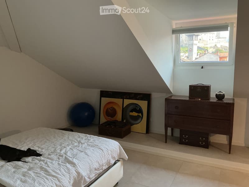 Cozy attic bedroom with slanted ceilings, a large window offering a city view, a bed with white bedding, a chest of drawers, a picture frame, and a large exercise ball.