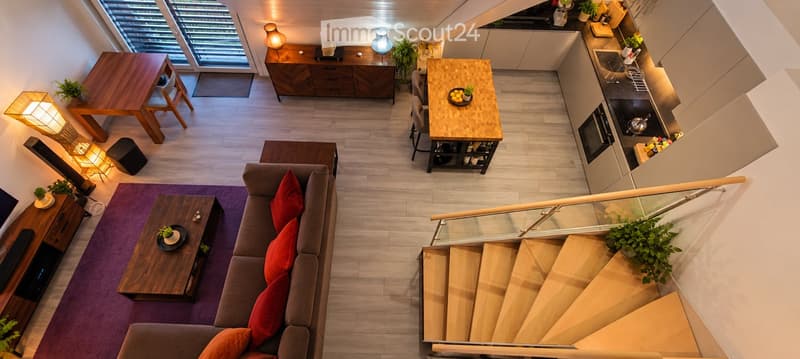 A modern, open-concept living and dining area with a grey sofa, wooden coffee table, dining table, and a kitchen area featuring a stove, sink, and refrigerator. The space includes a staircase leading to an upper level.