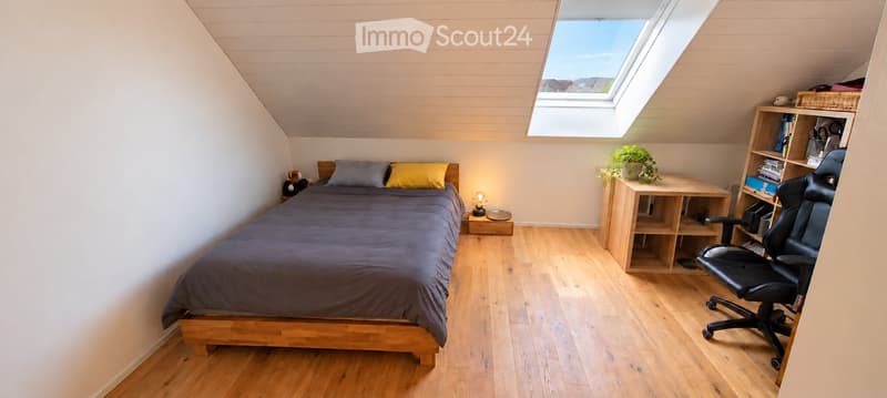 Cozy attic bedroom with wooden floors, slanted ceilings, skylight window, bed with grey duvet, nightstand with lamp, bookshelf, and small table with plant.