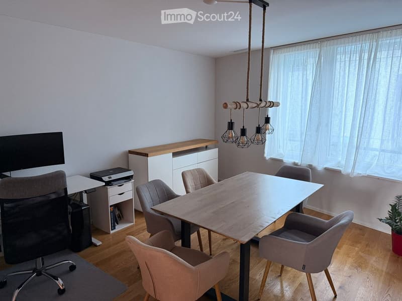 A modern, well-lit room with a large wooden table surrounded by chairs, a desk with a screen, and a cabinet with a printer. The room features wooden flooring and large windows with sheer curtains.