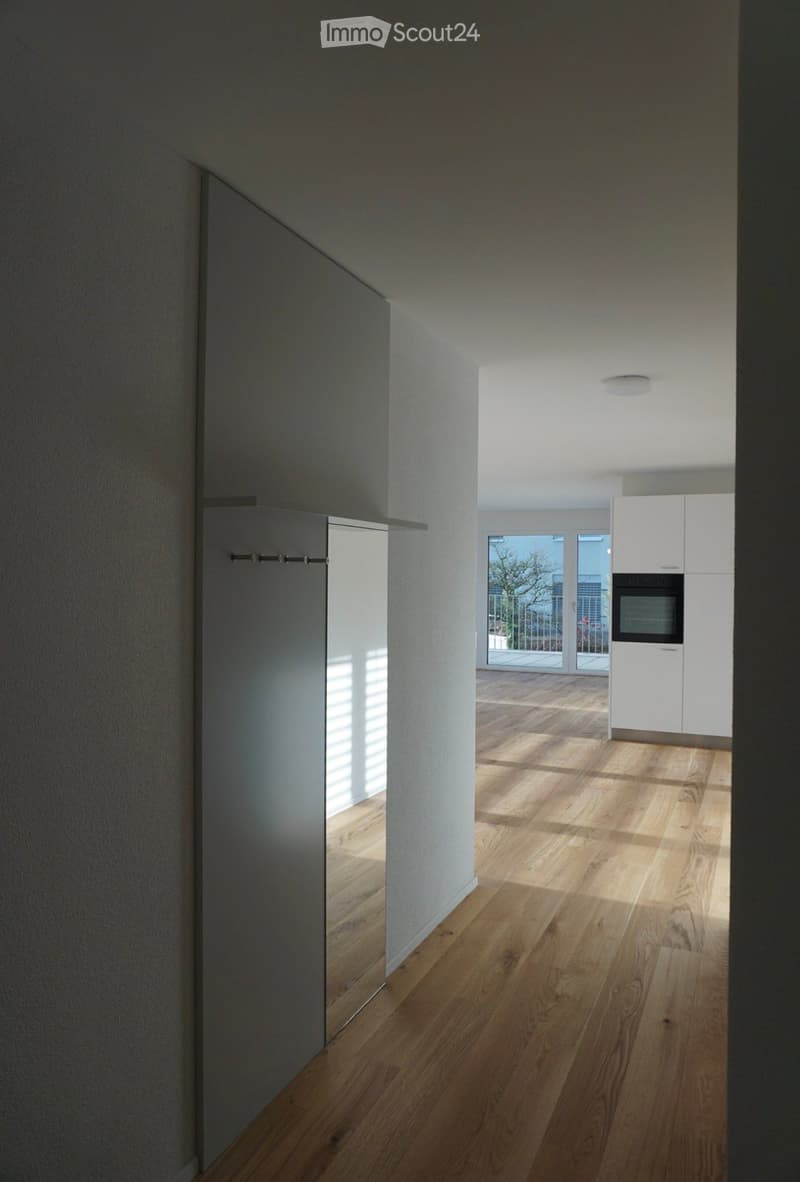 A modern, minimalist hallway with light wooden flooring, white walls, and a recessed ceiling light. The hallway leads to a kitchen area with white cabinets and a built-in oven, visible through an open doorway.