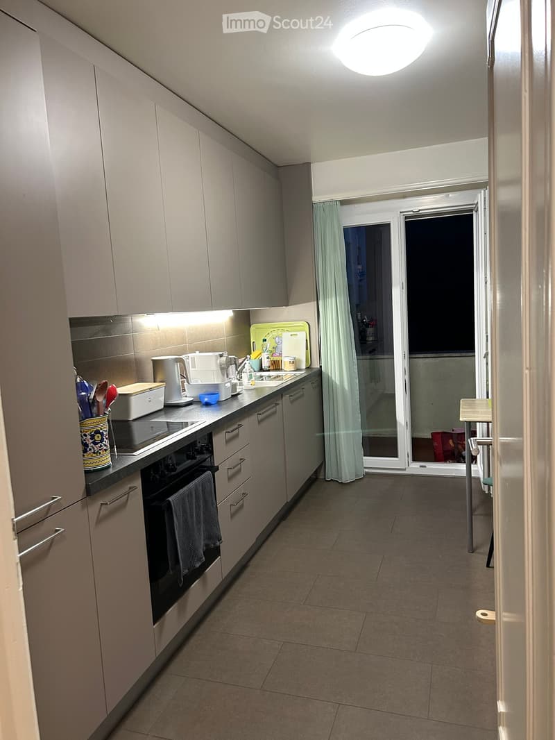 Modern kitchen with light grey cabinets, stainless steel appliances, and a tiled floor. The kitchen features an oven, coffee maker, and various utensils on the counter. A small table and chair are visible in the adjacent balcony area.