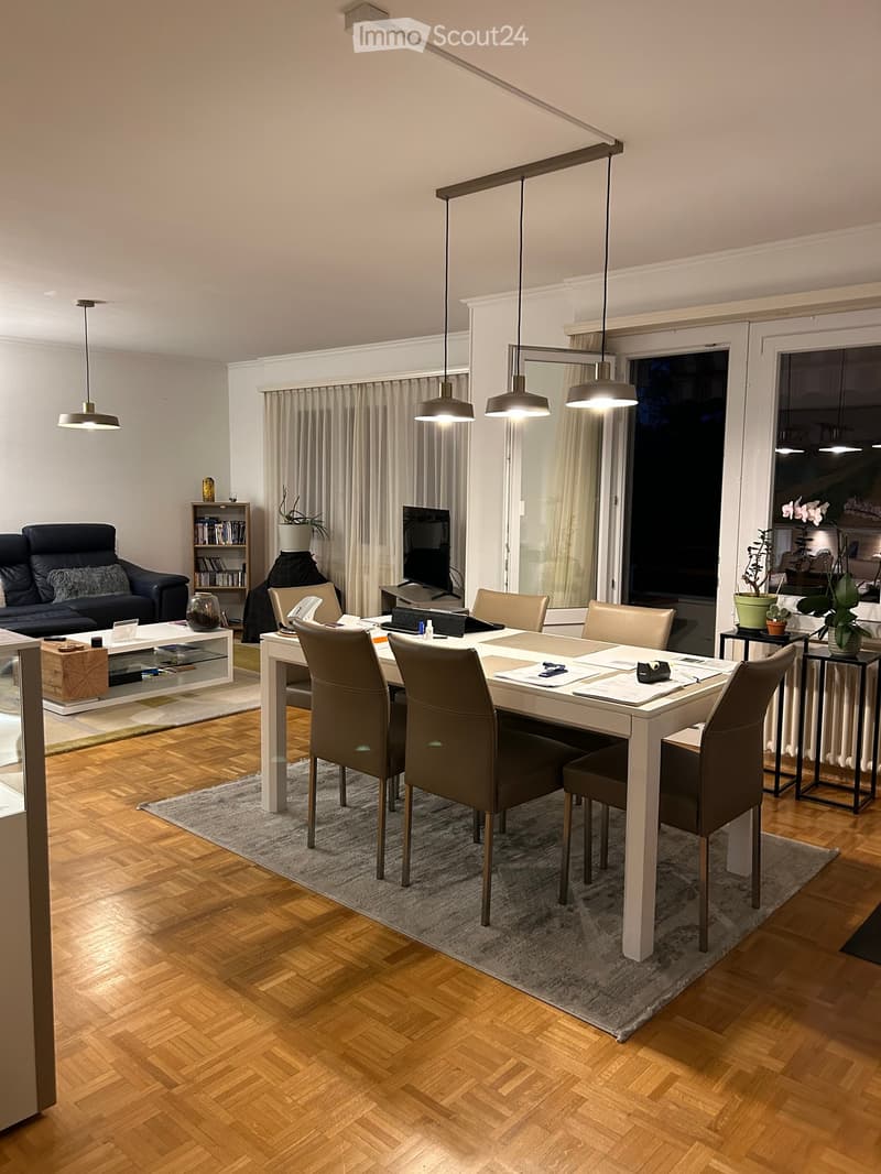 Modern living and dining area with wooden herringbone flooring, pendant lights, a sofa, coffee table, dining table with chairs, and a television on a stand.