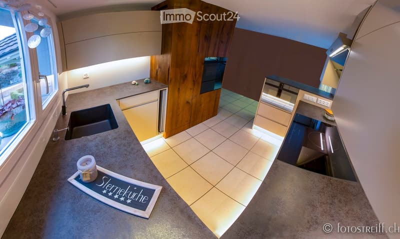 Modern kitchen with light wood cabinets, stainless steel sink, and a central island countertop. Features a candle and a decorative sign on the island.