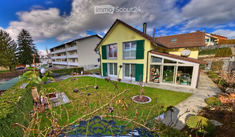 A two-story yellow house with green shutters, a well-maintained garden, and a small greenhouse attached to the side.
