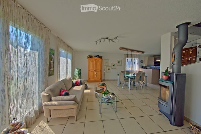 Bright living room with a beige sofa, glass coffee table, fireplace, dining area with a table and chairs, and a kitchen area in the background.