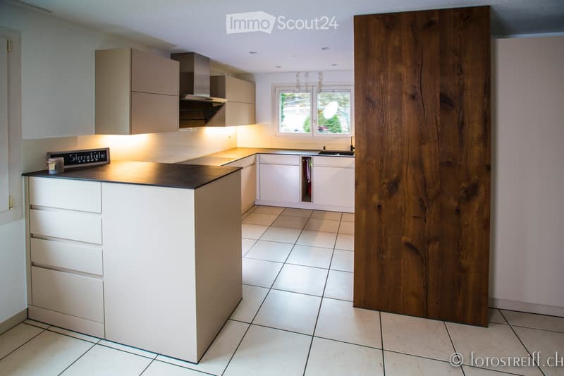 Modern kitchen with white cabinets, a central island, and a large window. Features include a range hood, sink, and a wooden panel accent wall.