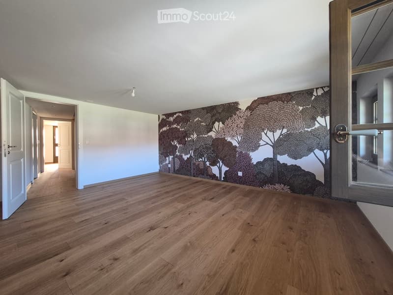 Empty room with wooden flooring, white walls, and a large mural of trees on one wall. The room has multiple doors and a window with a view of another space.