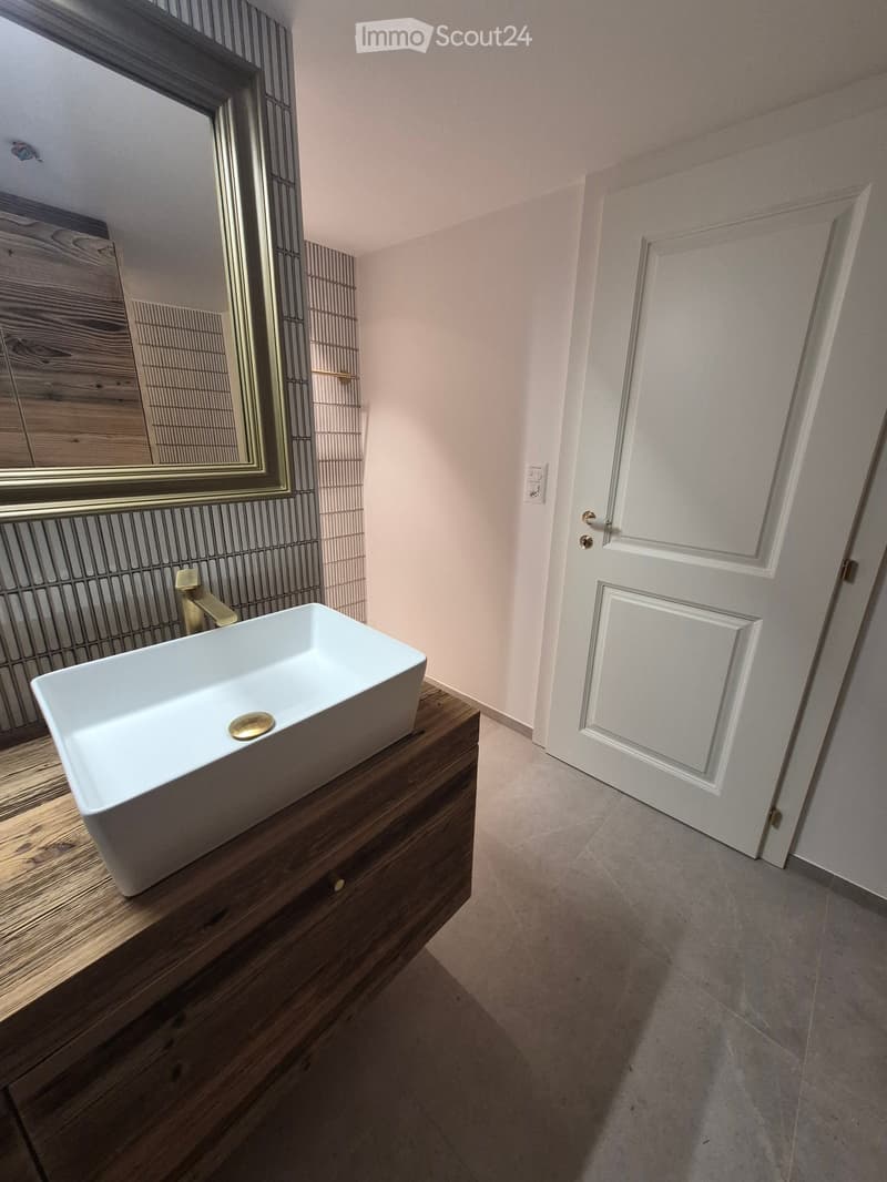 Modern bathroom with a white sink on a wooden vanity, a gold faucet, and a large mirror with a decorative frame. The room has light-colored walls and a white door.