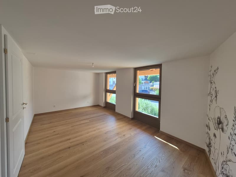 Empty room with wooden flooring, white walls, and large windows offering a view of the outside. The room has a door on the left and a wall with a floral design on the right.