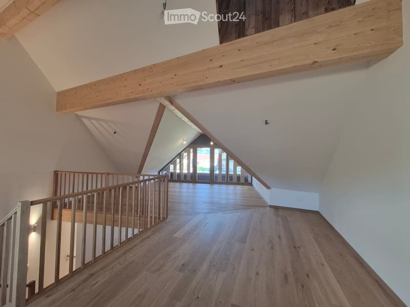 Spacious, open hallway with wooden flooring and exposed wooden beams. Features a wooden staircase with a railing leading to an upper level, and large windows at the end allowing natural light to flood the space.