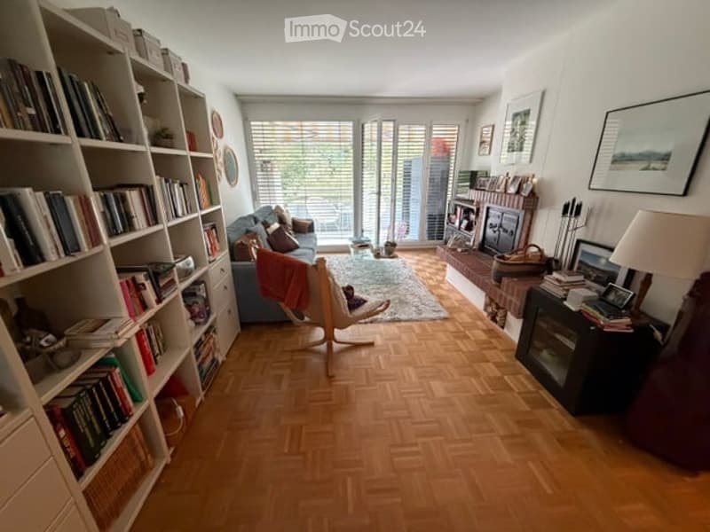 Living room with wooden parquet flooring, large windows, balcony access, sofa, bookshelf, chair, table lamp, fireplace, and picture frames.