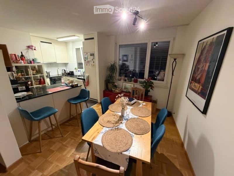 Open-plan living and dining area with modern furniture, including a dining table with six chairs, a kitchen counter with stools, and shelves with various items. The space features wooden flooring, white walls, and large windows with blinds.