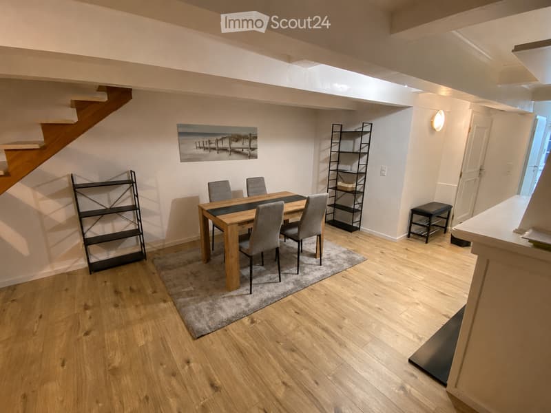 Dining area with wooden dining table, four chairs, two black shelves, a small stool, and a painting on the wall. Wooden flooring and a gray rug under the table.
