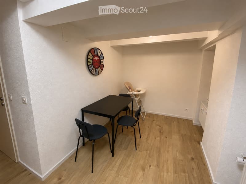 A small, furnished room with a black table, four chairs, a baby chair, and a wall-mounted clock. The room has light wooden flooring and white walls.