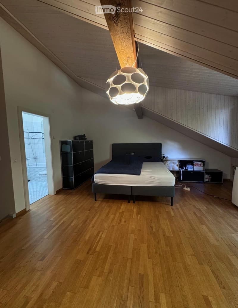 Modern attic bedroom with wooden floors, slanted wooden ceiling, and built-in storage. Features a double bed, wardrobe, nightstand, and a unique hanging lamp.