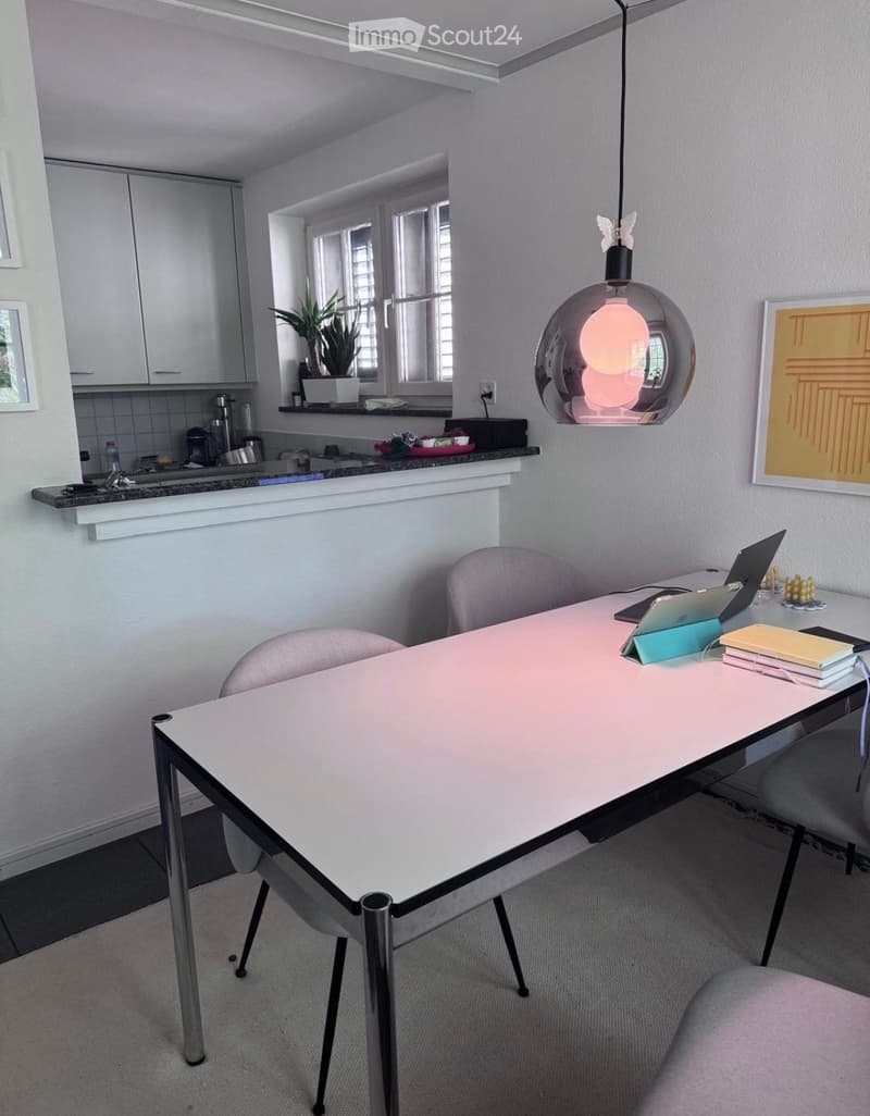A modern, well-lit room with a white table and chairs, a laptop, a kitchen area with cabinets and a sink, and a hanging light fixture.