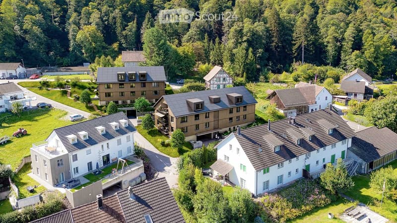 Aerial view of a residential area with multiple houses, surrounded by greenery and trees. Houses have varied architectural styles, some with balconies and garages.