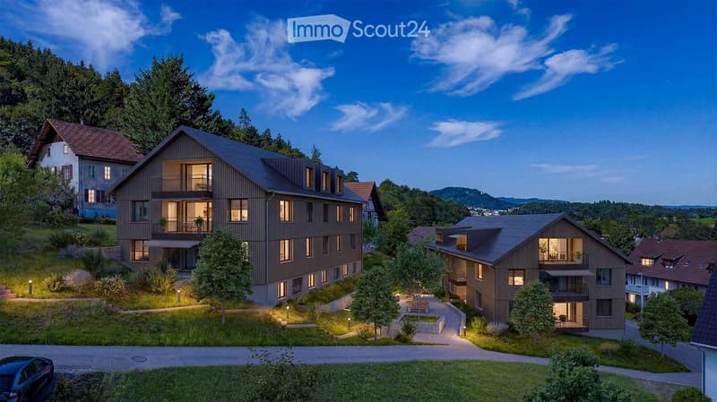 A modern residential complex with multiple buildings, balconies, and surrounding greenery, set in a hilly landscape with trees and a clear sky.