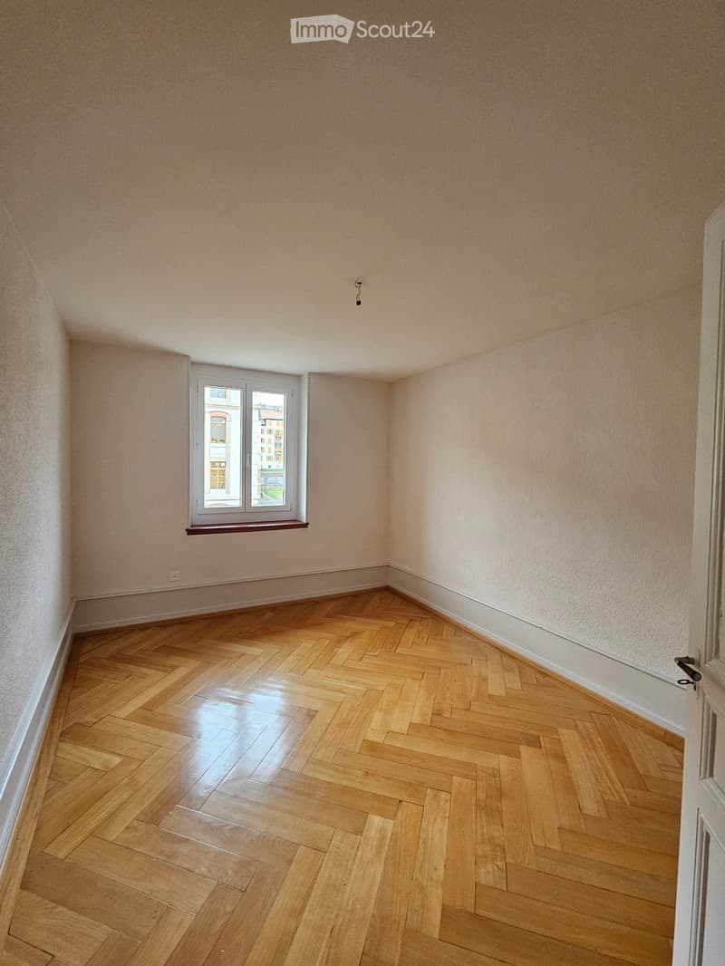 Empty room with light-colored walls and polished wooden herringbone flooring, featuring a single window with a small sill and a door frame visible on the right side.