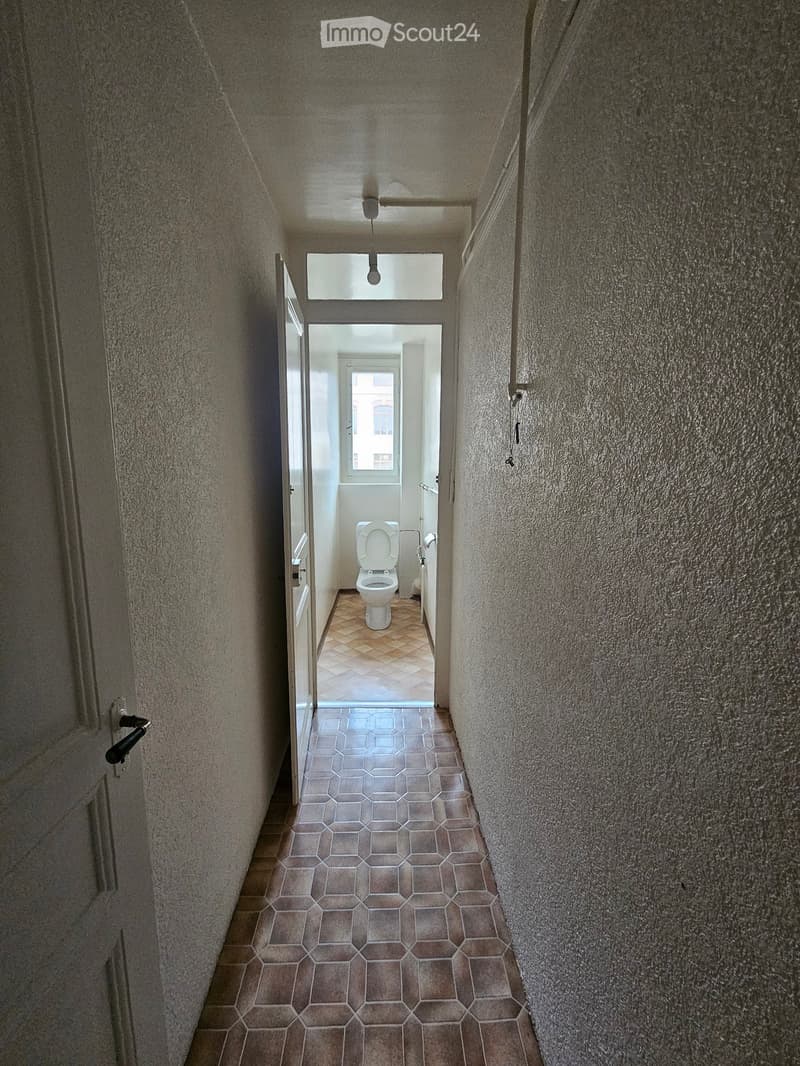 A narrow hallway with tiled flooring and textured walls, leading to a bathroom with a toilet and a window.