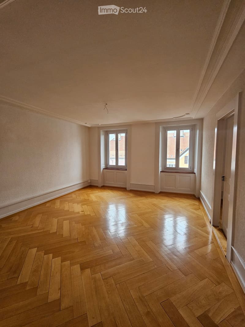Empty room with wooden herringbone flooring, light-colored walls, and two windows with radiators beneath them.