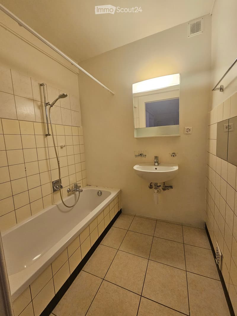 Modern bathroom with a bathtub, sink, mirror, and showerhead. Light-colored tiles and a sleek design.