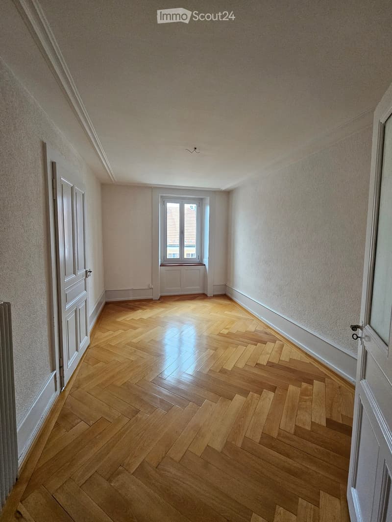 Empty room with wooden herringbone flooring, light-colored walls, and a window with a radiator below it. The room has a door on the left side and a small light fixture on the ceiling.