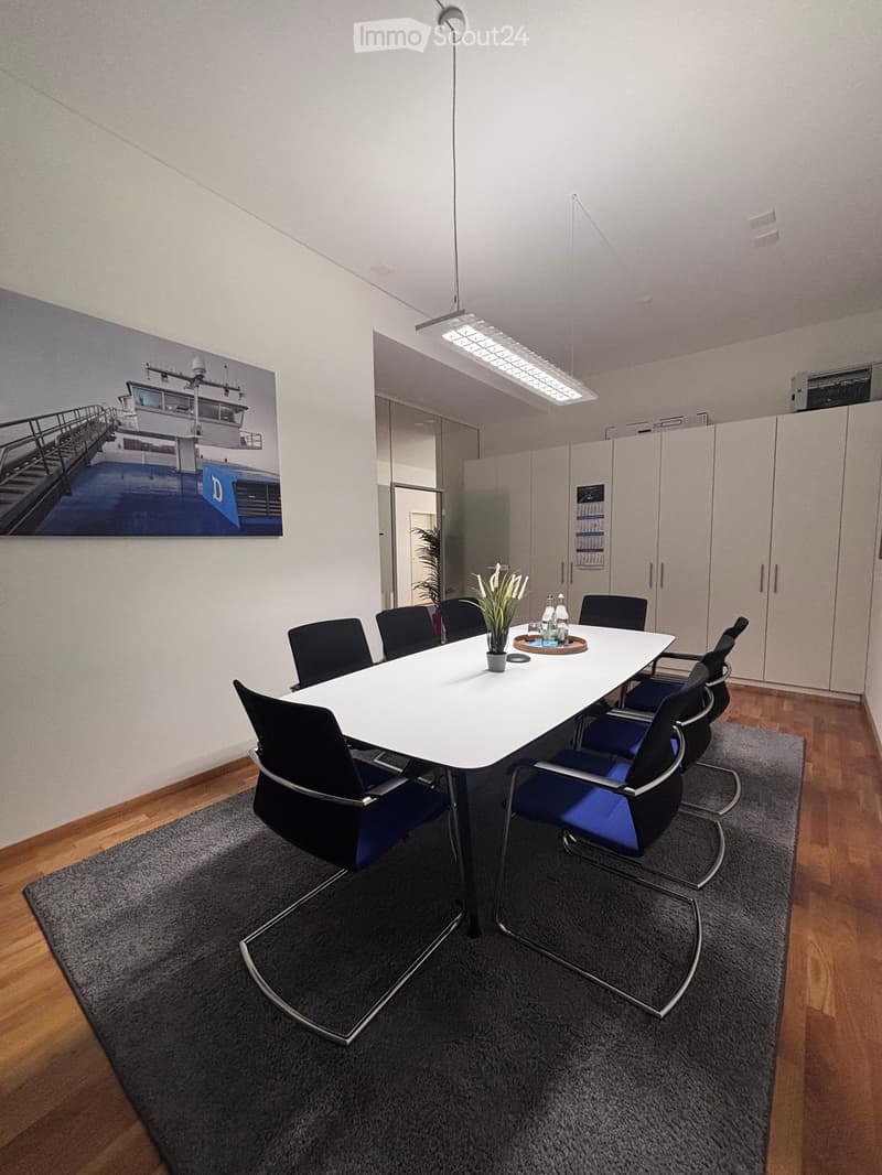A conference room with a long white table, black and blue chairs, a vase with flowers, and a potted plant on the table.