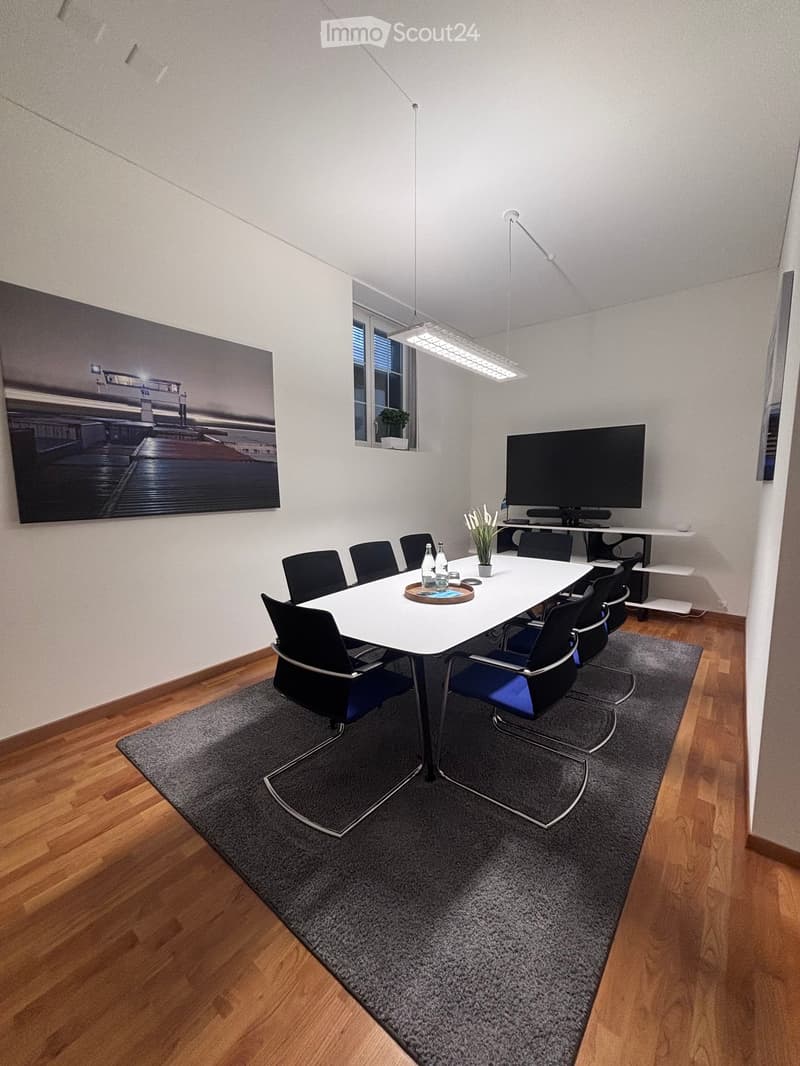 A conference room with a long white table, black chairs, a TV on a shelf, a potted plant, and a vase on the table.