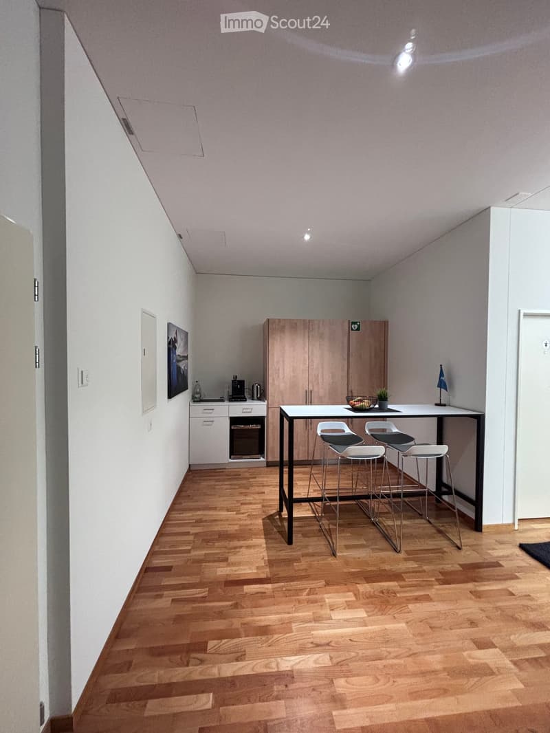 A modern, minimalist kitchen with wooden floors, white walls, and a sleek table and chairs. The kitchen includes a cabinet, a plant, and a flag. There is a basket of fruits on the table, a coffee machine, a kettle, and a bottle on the counter. A picture i
