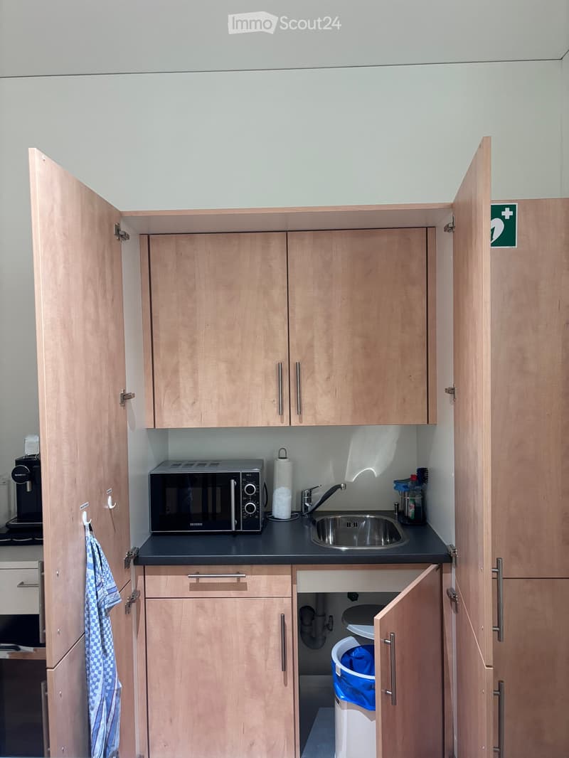 A small kitchen area with wooden cabinets, a black countertop, a microwave, sink, and a trash can.