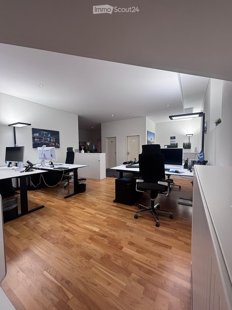 Modern office with wooden floors, white walls, and two desks equipped with computers, chairs, and various office equipment.