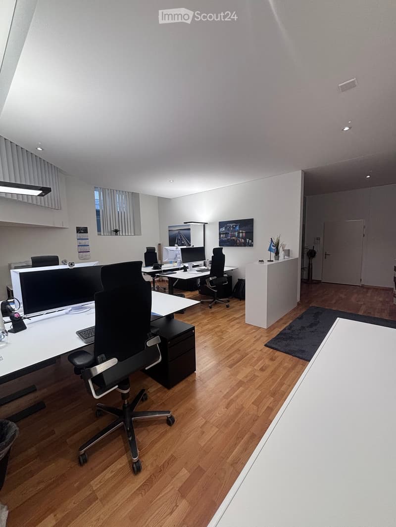 Modern office space with desks, chairs, and computer monitors. White walls and wooden floors.